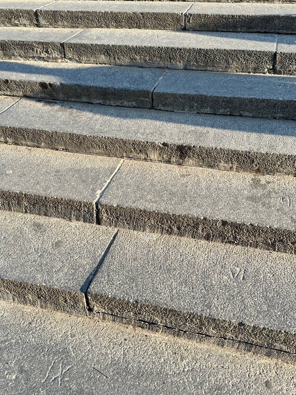 Gros plan sur des marches de pierre grise aux bords rugueux, avec de fins joints entre les dalles. Des ombres nettes couvrent les marches du haut ; un chiffre romain VI est gravé sur une dal