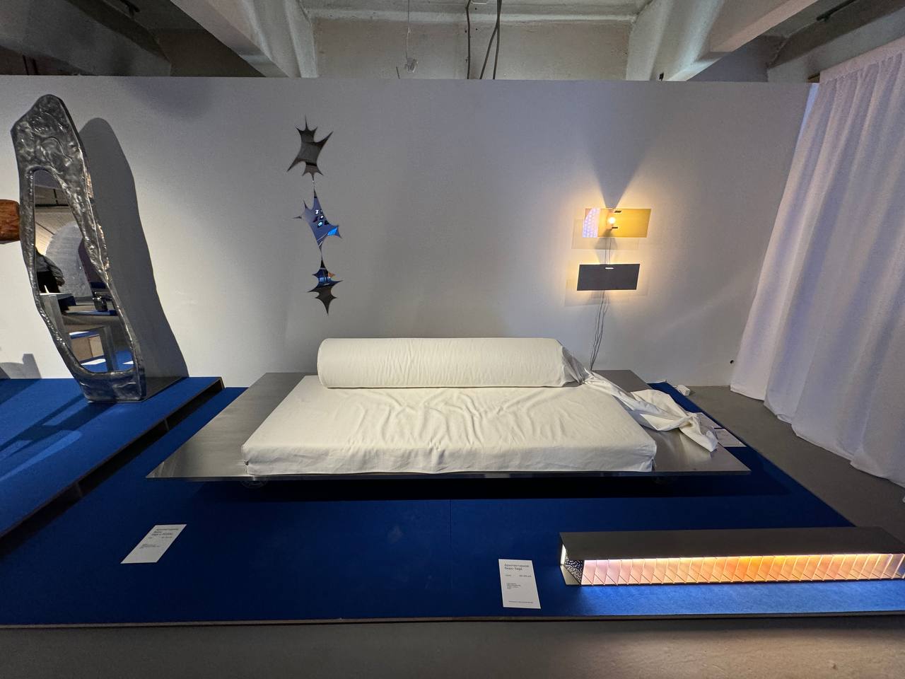 Sur un podium bleu, un canapé à matelas blanc et traversin repose sur une plateforme en acier. Autour : un miroir en métal coulé, une applique et un panneau lumineux.