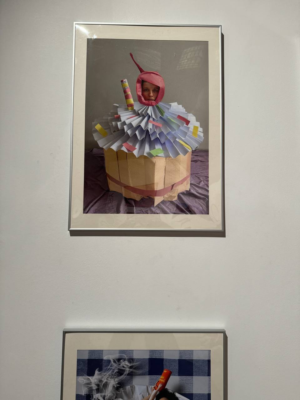 Sur un mur blanc, une photo encadrée montre une personne en cupcake de papier, coiffée d’une cerise rose. En dessous, une autre œuvre : tissu à carreaux et fumée.