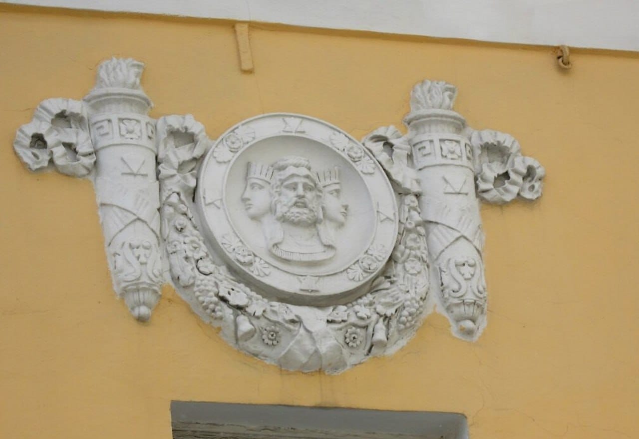 Médaillon en stuc blanc : une tête barbue au centre entre deux profils couronnés. Une guirlande de fleurs et de fruits l'entoure, avec deux torches ornées de rubans, sur un mur jaune.