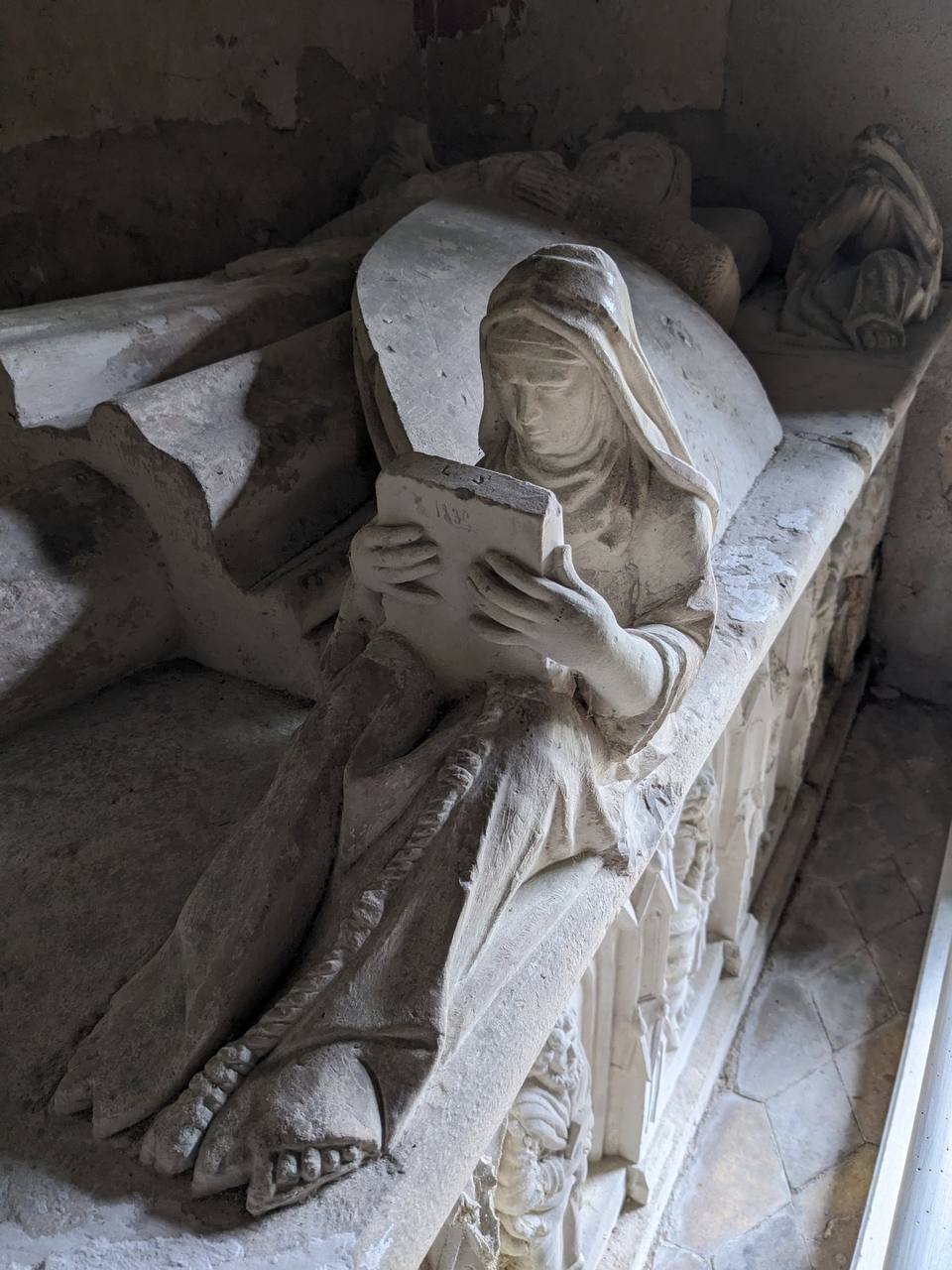 Statuette de pierre d'une femme voilée tenant un livre, assise au bord d'un tombeau orné de niches gothiques.