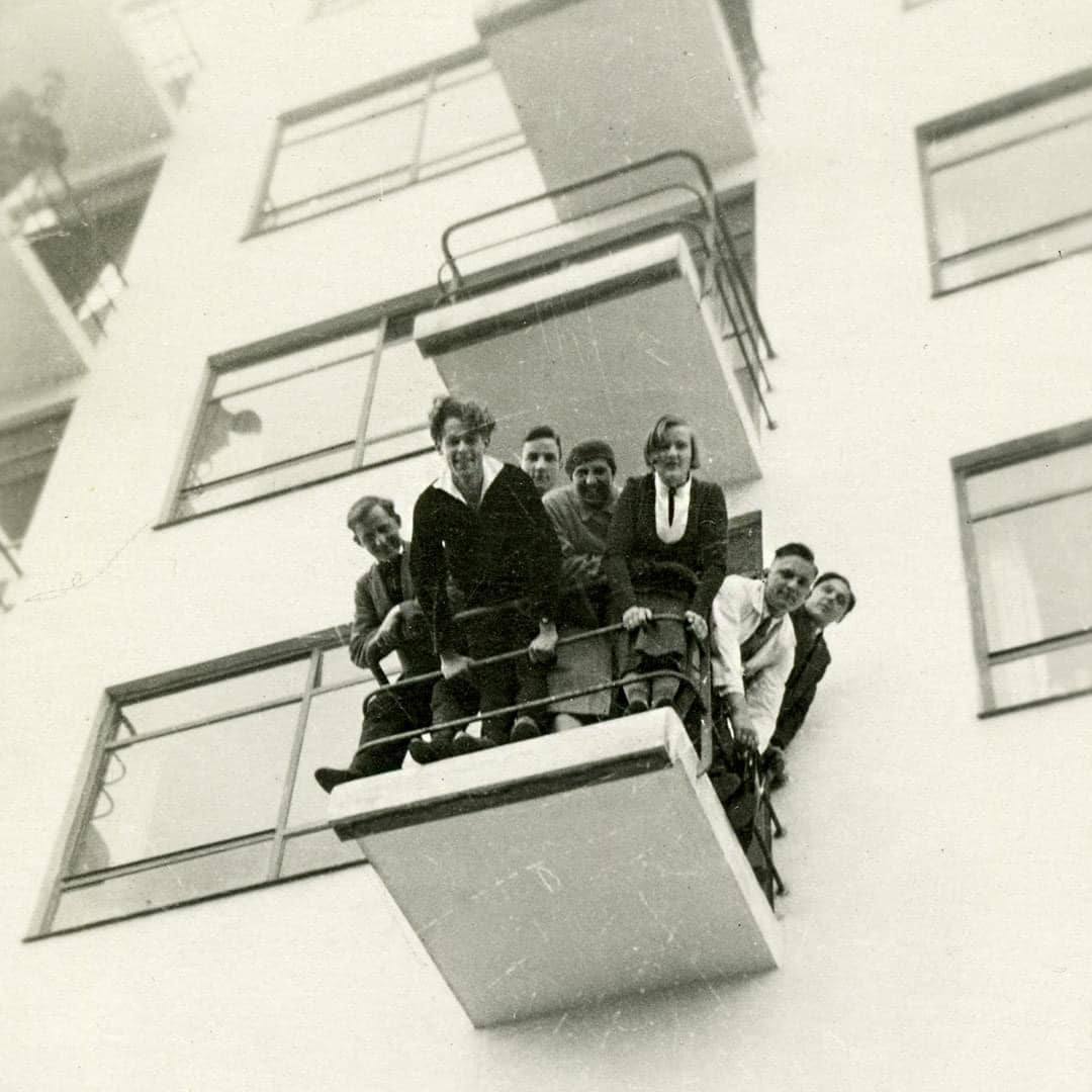 Photographie en noir et blanc d’un groupe de personnes regardant vers le bas depuis le balcon d’un immeuble blanc aux grandes fenêtres.