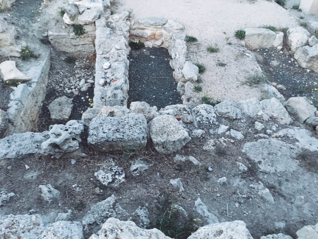 Stone remains of a foundation with three graves, the middle one filled with dark soil, surrounded by rubble and sparse grass.