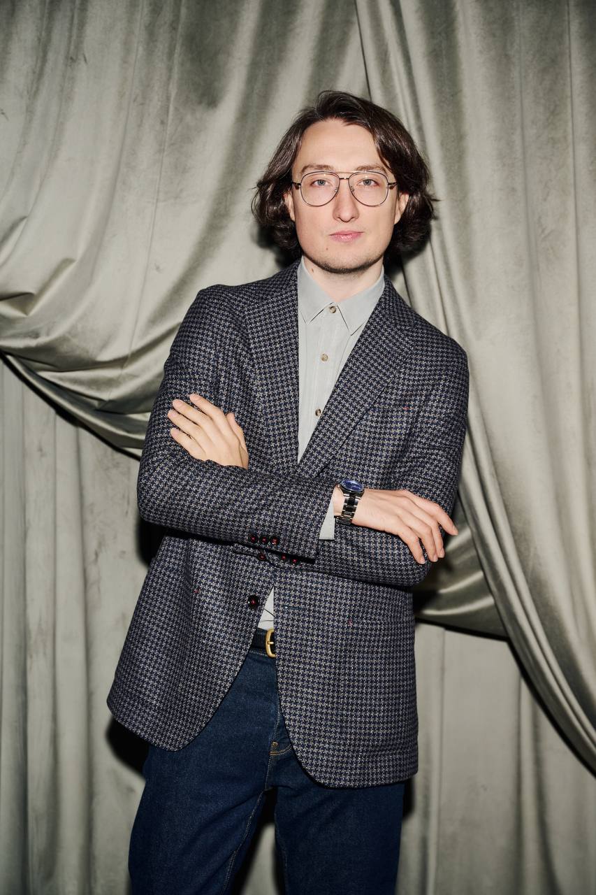 Un jeune homme à lunettes, aux cheveux ondulés foncés, en blazer à carreaux et jean, bras croisés devant un rideau drapé gris-vert.