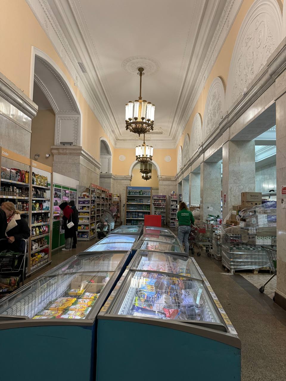 A shop hall with a stucco ceiling, chandeliers, arches, freezer cases and shoppers among shelves of goods.