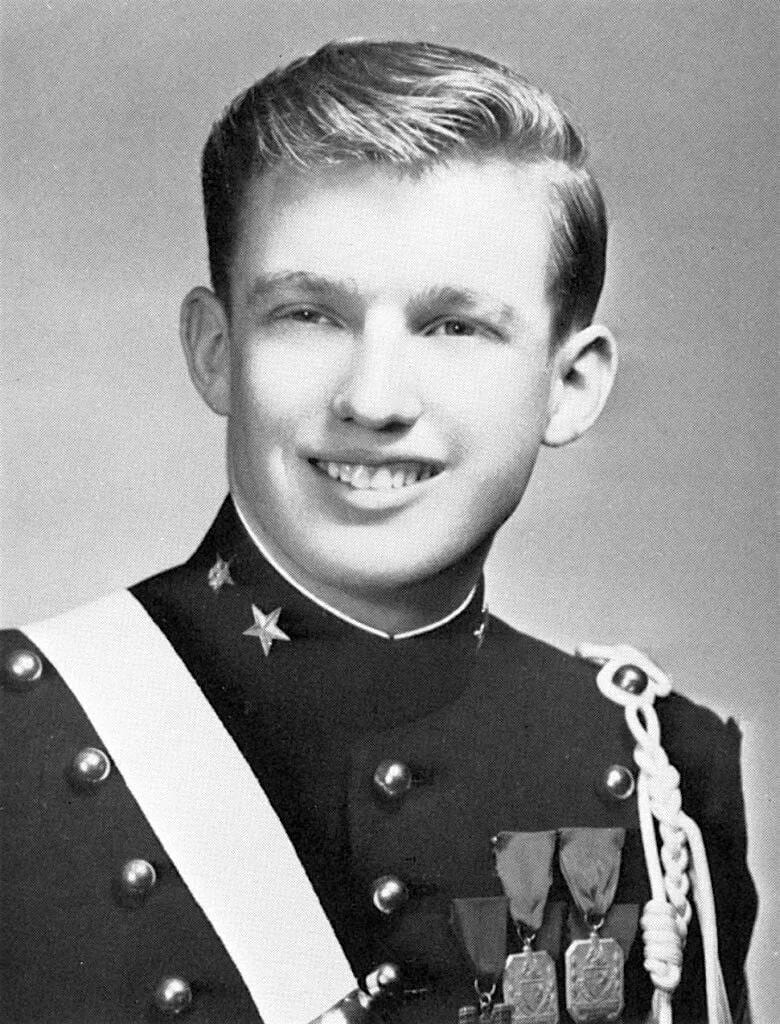 Black-and-white portrait of a smiling young man in a dark uniform with a white sash, buttons, medals and a braided cord.