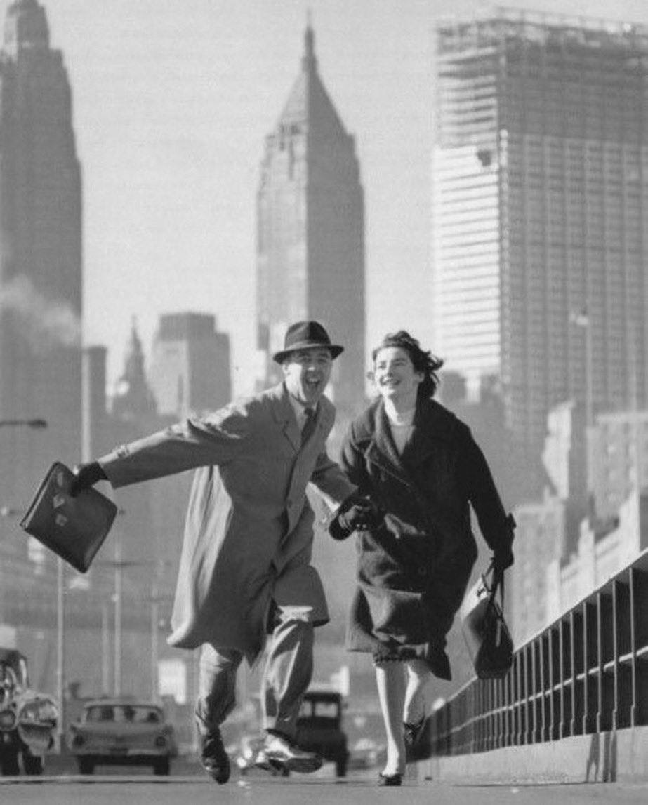 Black-and-white photograph of a man in a hat with a briefcase and a woman in a coat running hand in hand along a street with skyscrapers behind them.