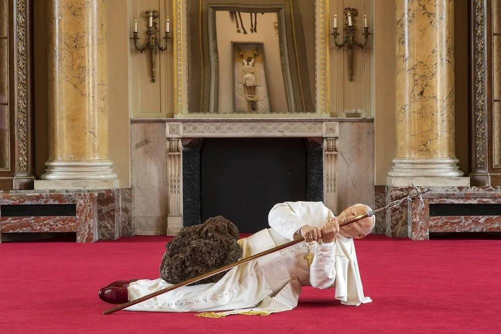 A figure in white papal vestments lies on a red carpet in an ornate hall with marble columns and a fireplace, gripping a staff while pinned beneath a dark rock.