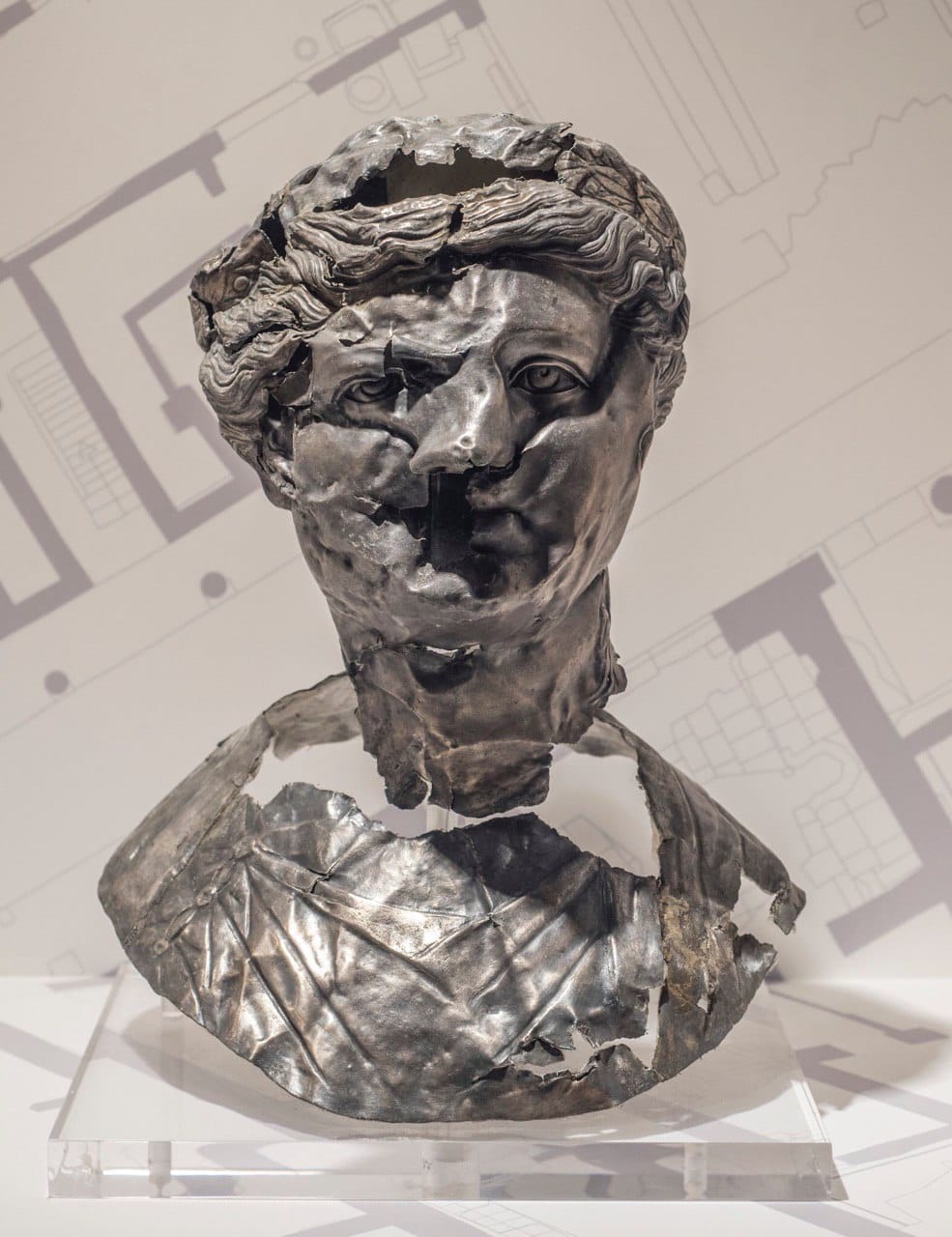 A damaged silver bust with wavy hair and a deformed face stands on a transparent base against a background of architectural plans.