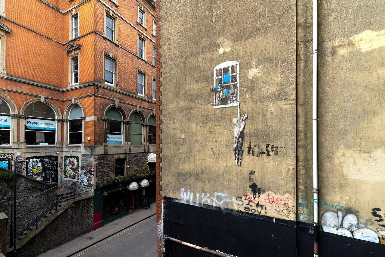 Mur d'un bâtiment de Bristol avec le graffiti de Banksy représentant un homme suspendu à une fenêtre, près d'un immeuble de briques orange.