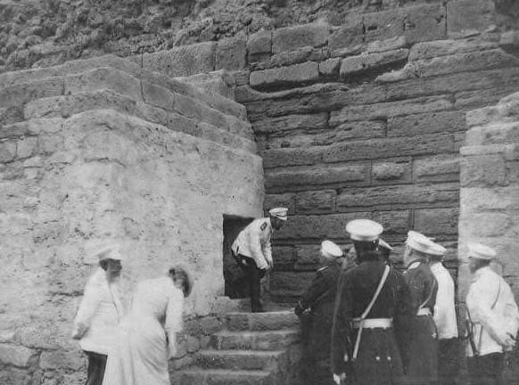 Black and white photograph of men in white caps and women by a stone wall with steps and a dark doorway, one man stepping into the opening.