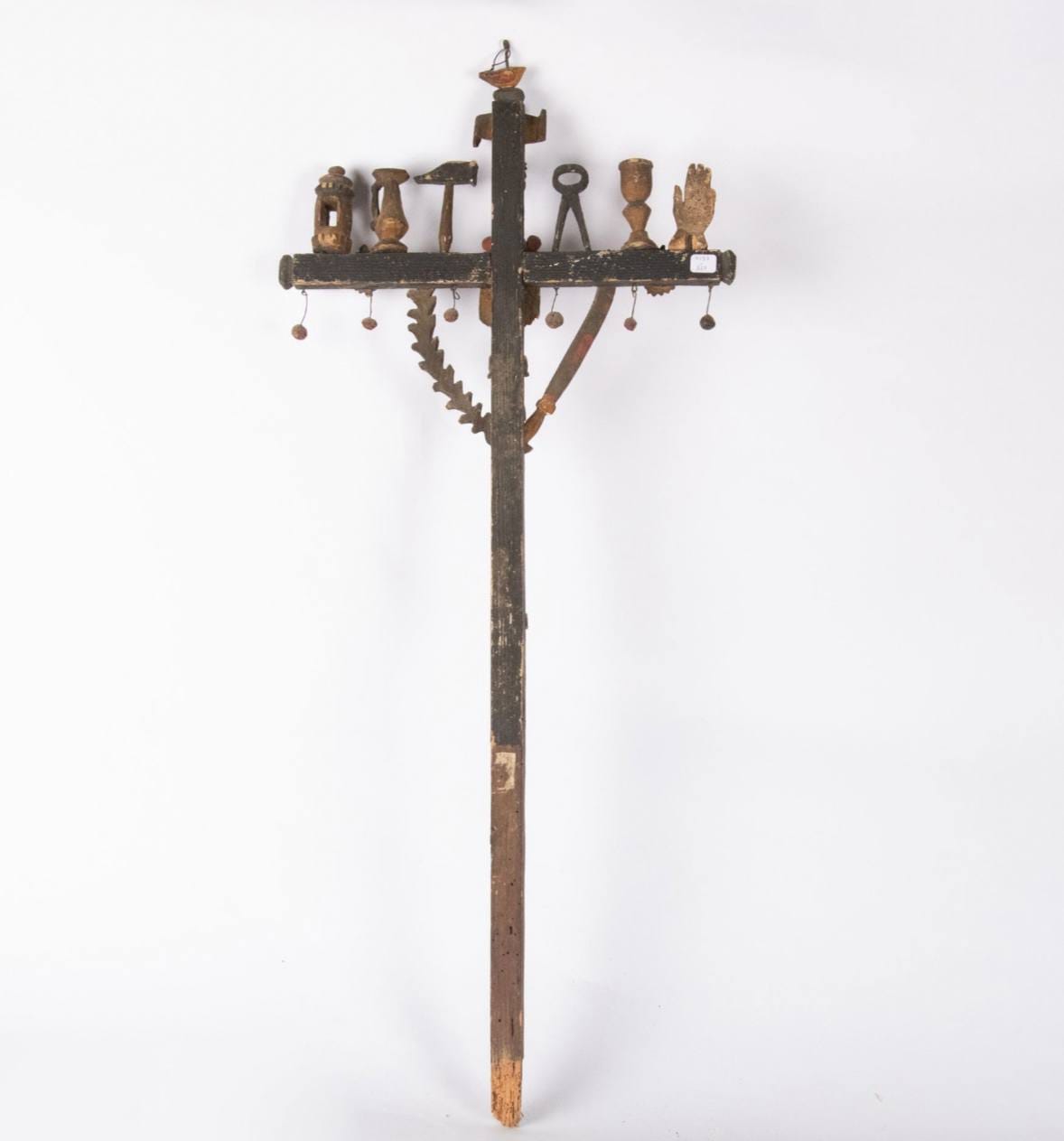 Croix en bois sombre à la peinture usée. Sur la traverse sont posés des objets sculptés : lanterne, cruche, marteau, tenaille, coupe, main. Des boules pendent dessous ; une scie et une courb