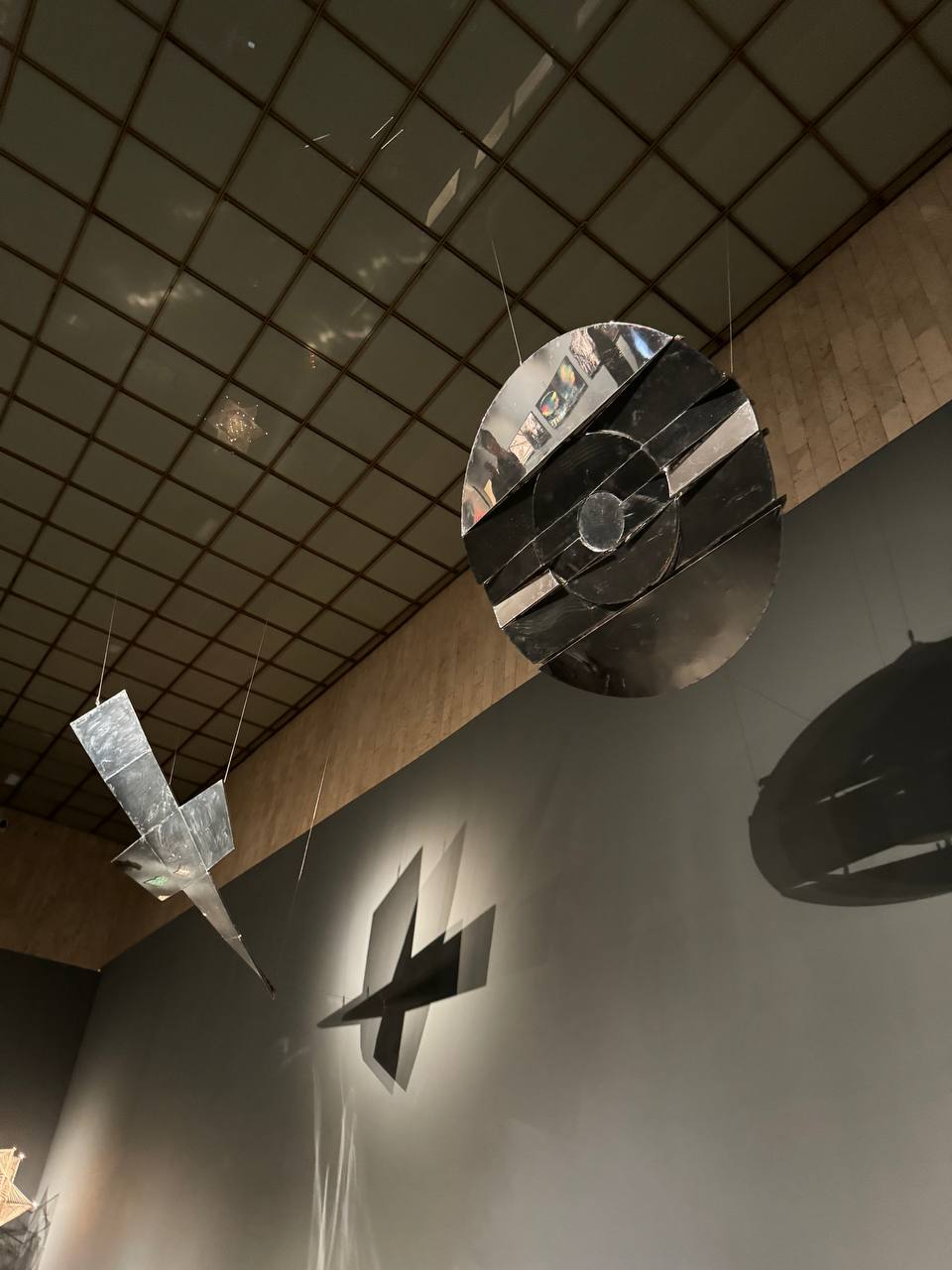 Under a coffered museum ceiling, a mirrored disc and an angular metal form hang on thin wires; their shadows fall on the grey wall.