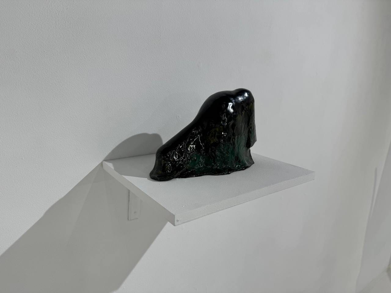 A small black ceramic sculpture with glossy glaze and greenish tones sits on a white shelf against a white wall, casting a sharp shadow beside it.