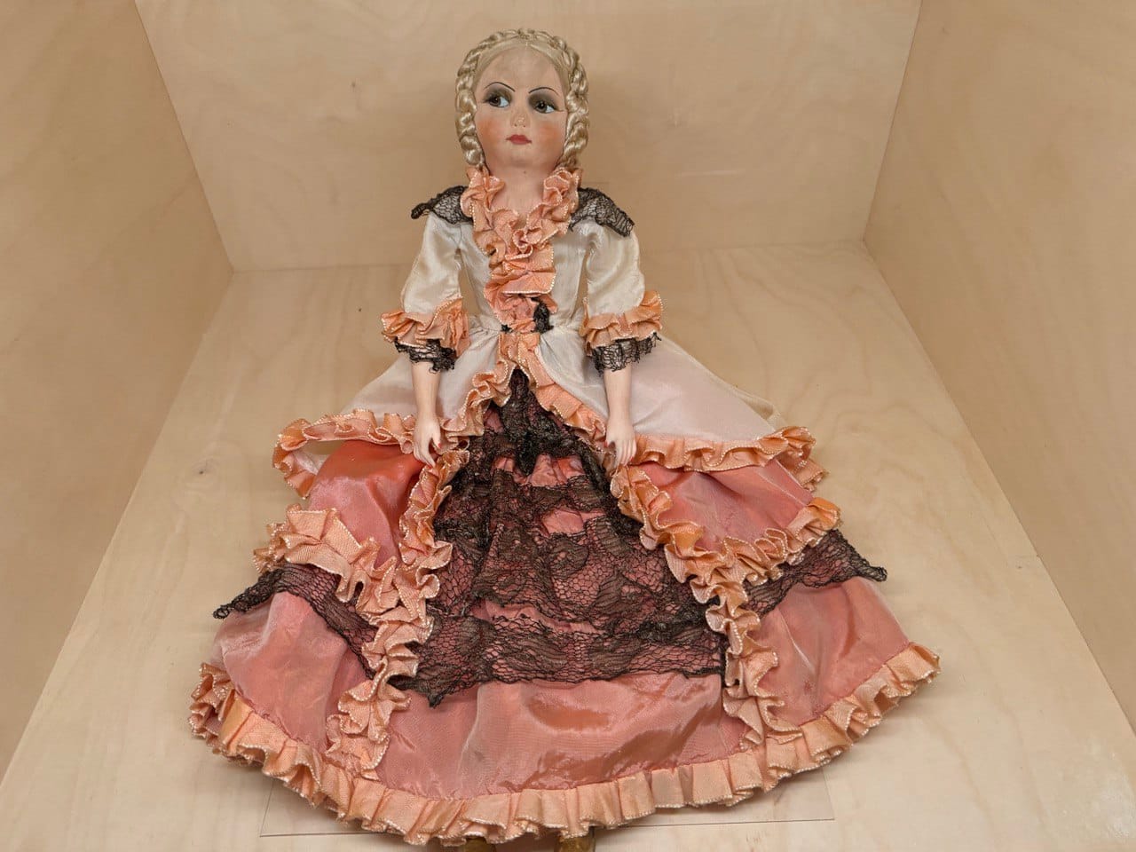 A doll with blond braids and painted eyes sits in a wooden box. She wears an 18th-century-style satin dress with peach ruffles, a pink skirt and black lace.