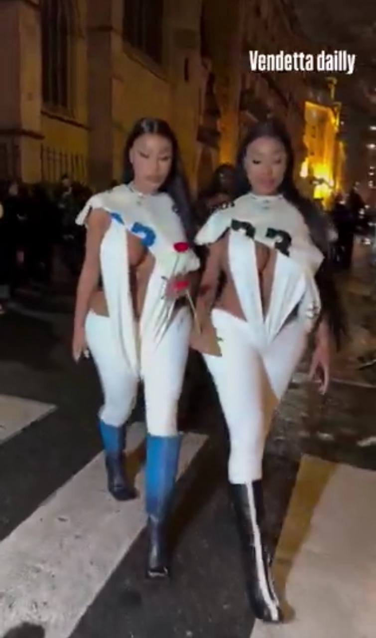 Two women with long dark hair, in white outfits with numbers and tall boots, cross a crosswalk at night. One holds a rose; buildings and a crowd are behind them.