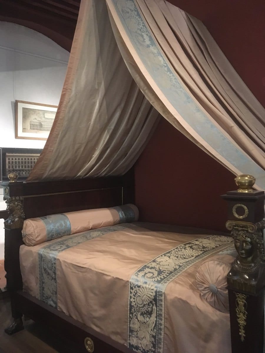 Wooden bed with a pink silk canopy, patterned blue trim and bronze female heads on the posts, displayed in a museum room.