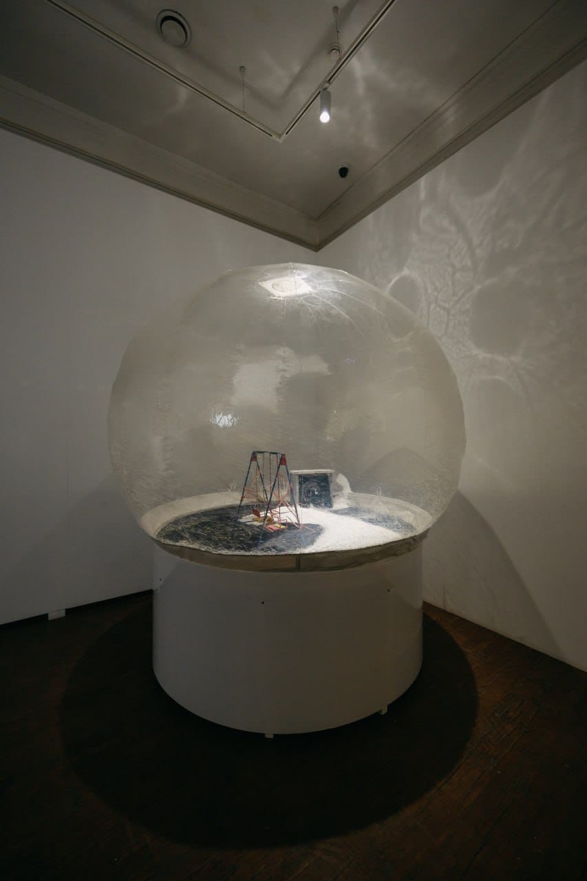 Un dôme transparent de la taille d'une pièce sur un socle blanc. À l'intérieur, des balançoires miniatures et un petit écran sur de la neige; des reflets de lumière couvrent les murs.