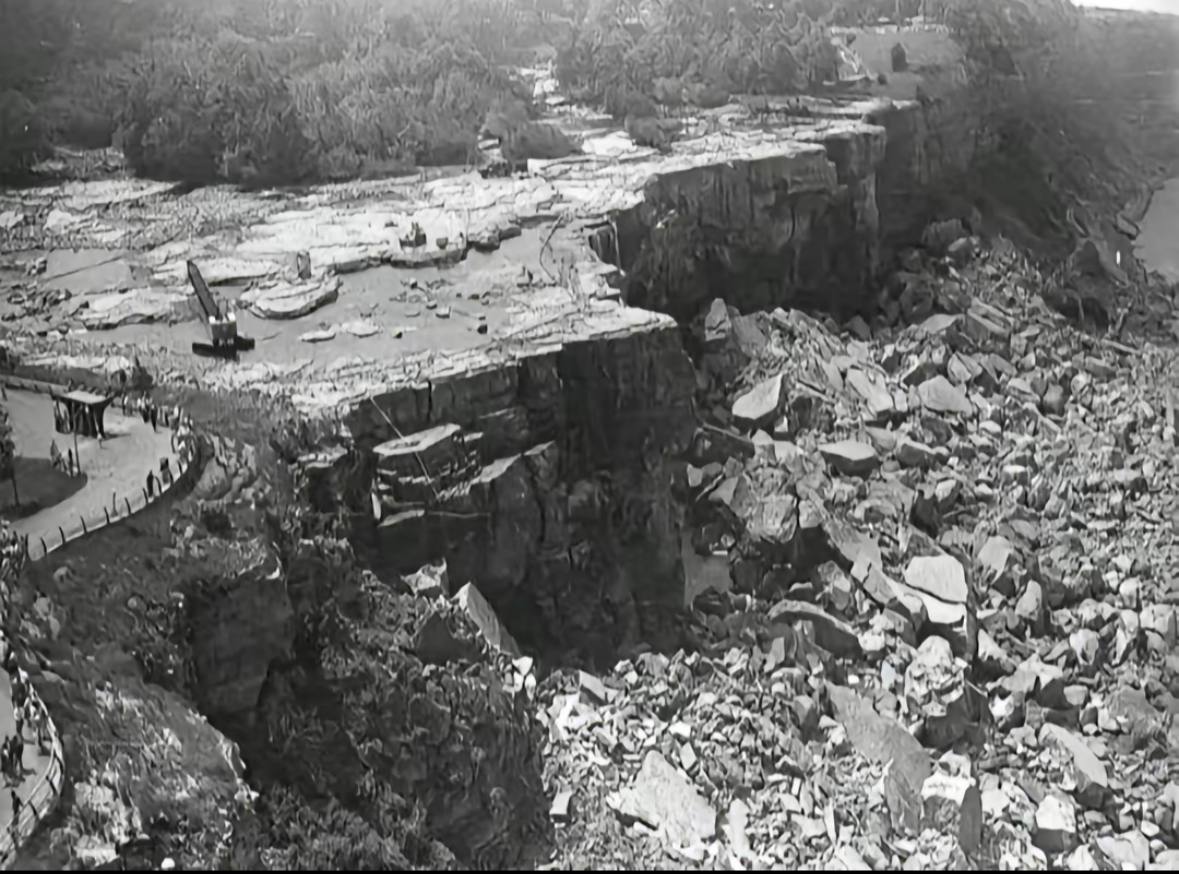 Photographie en noir et blanc d'un lit de rivière à sec, avec des corniches rocheuses, des amas de pierres brisées et un chemin clôturé à gauche.