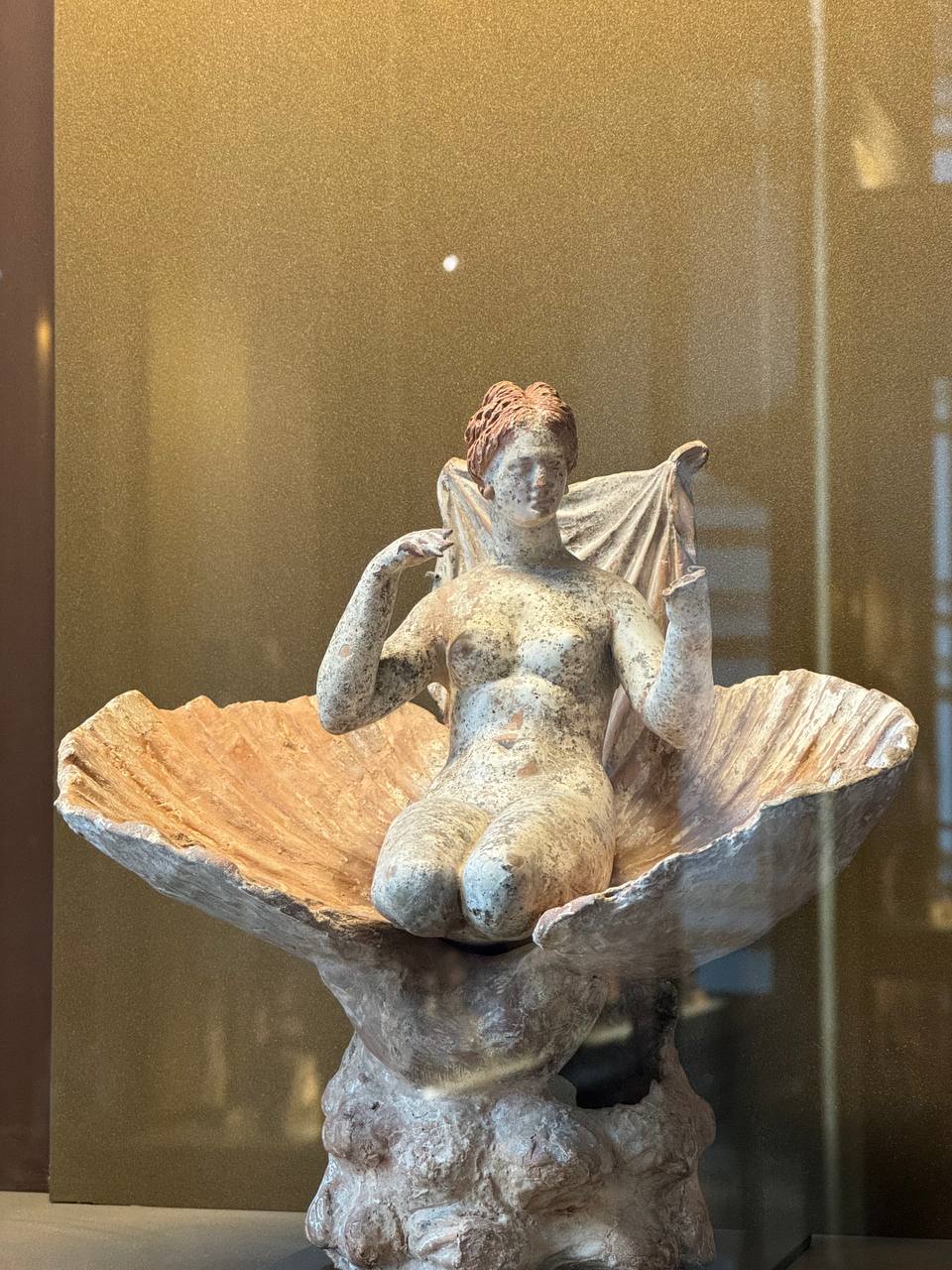 Statuette of a nude woman with reddish hair seated in an open shell, in a museum display case.