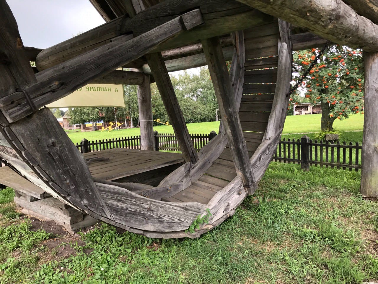 A massive wooden structure with curved beams on the grass, with a low picket fence and a rowan tree with red berries behind it.