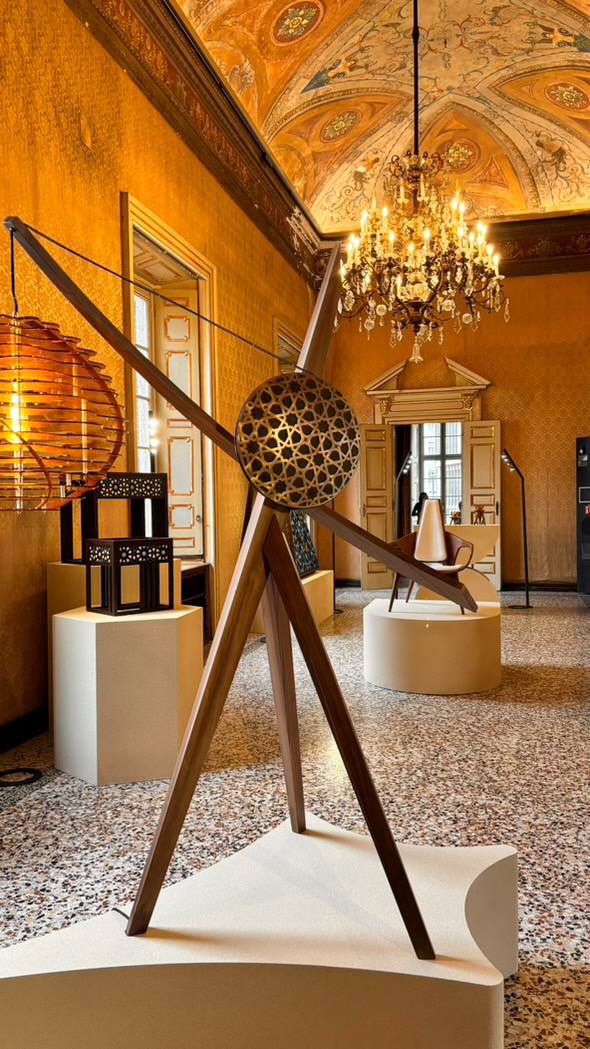 A wooden tripod sculpture with a perforated brass disc stands in a hall with golden walls, a chandelier and a frescoed vault. Chairs and a lamp sit on pedestals.