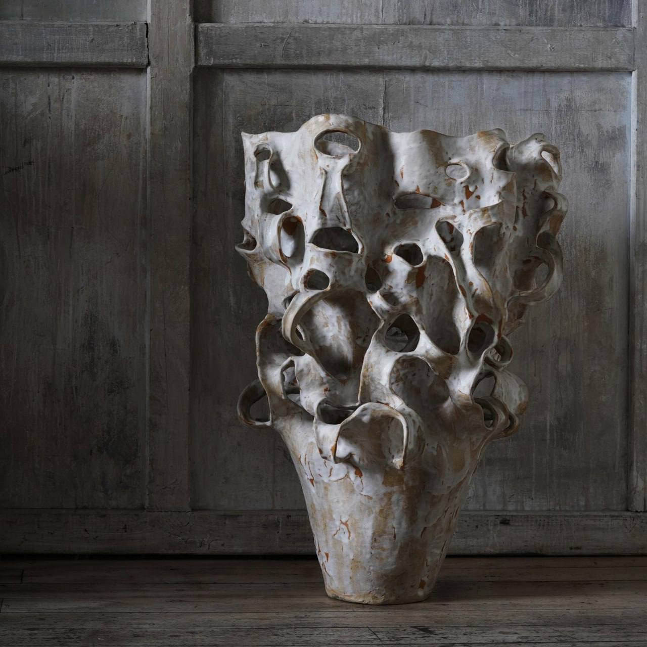 A tall white glazed vase-like sculpture with wavy edges and many openings stands on a wooden floor before a grey wall of weathered planks.