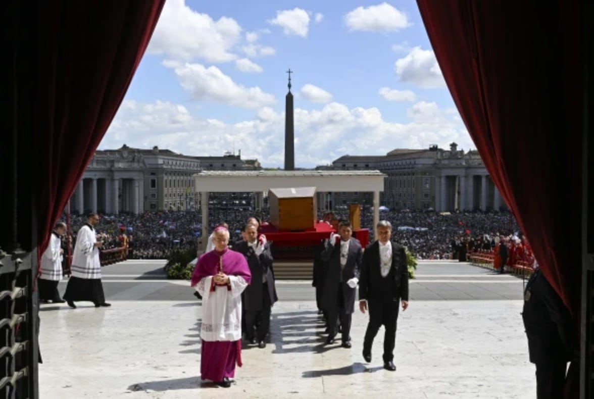 Un prélat en tenue violette précède des hommes en costume sombre portant un cercueil en bois. Au-delà des rideaux rouges, l'obélisque et une foule immense sur la place.