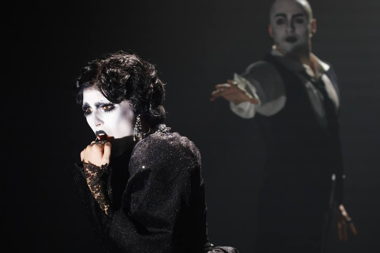 A woman with dark curls, in a black dress and lace glove, presses her fist to her lips. Behind her, blurred, a bald man in a waistcoat reaches out. Both wear white face paint.