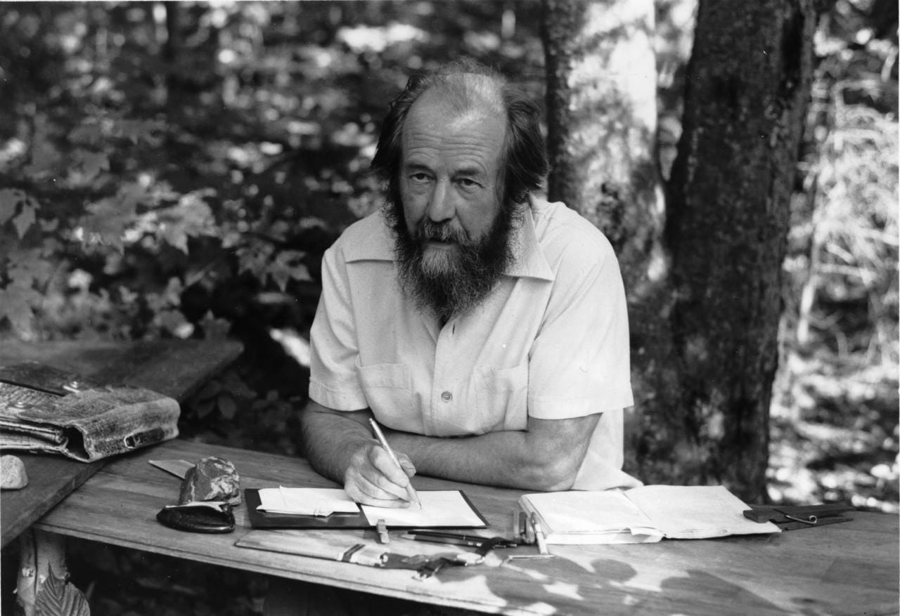 Black-and-white photograph of a bearded man in a short-sleeved shirt writing at a wooden table outdoors among trees.