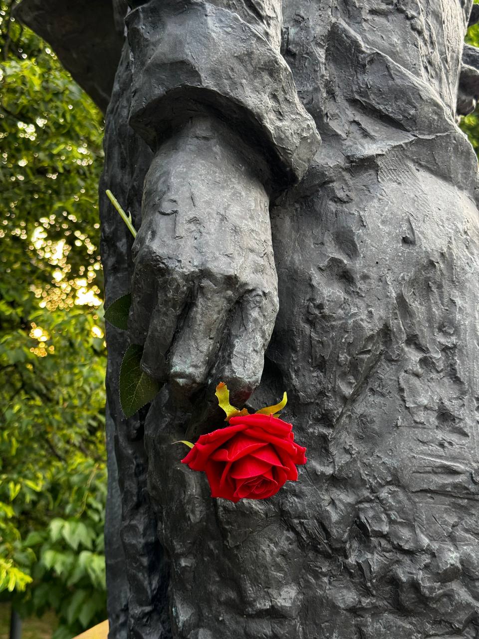 Gros plan sur la main et la manche d'une statue en bronze sombre, avec une rose rouge glissée entre les doigts et le corps. Feuillage vert flou en arrière-plan.