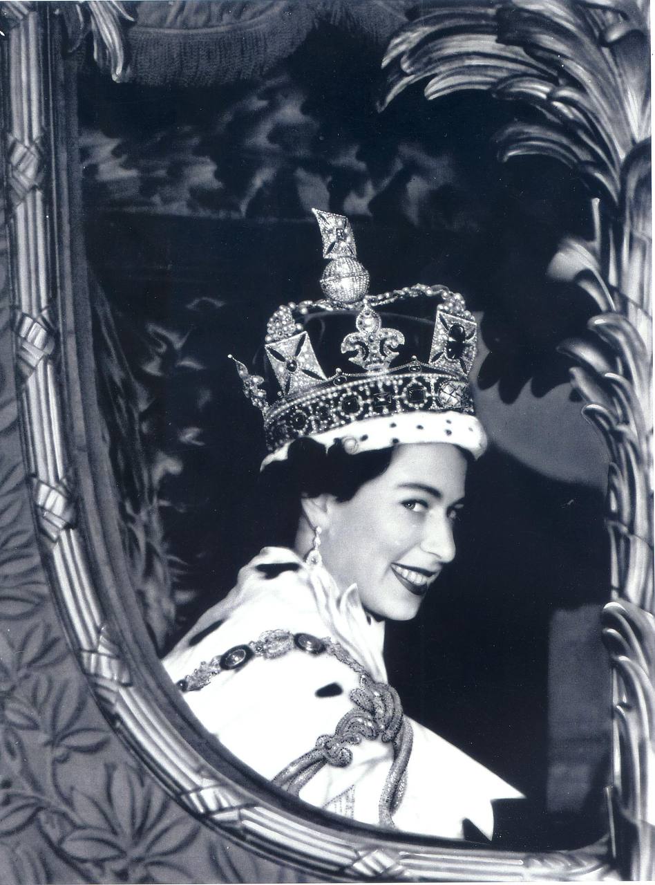 Photographie en noir et blanc d'une femme souriante portant couronne ornée de joyaux, manteau bordé d'hermine et pendants d'oreilles, se retournant dans un cadre sculpté.