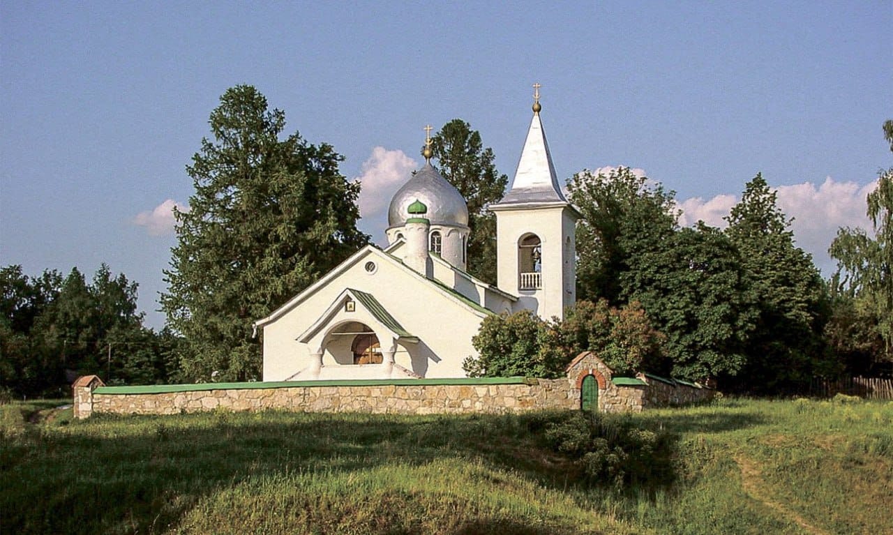 Une église blanche à coupole argentée et clocher, derrière un muret de pierre, entre des arbres et une prairie verte.