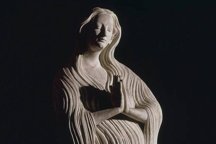 Sculpture en pierre de Marie-Madeleine aux longs cheveux et aux mains jointes en prière, sur fond sombre.