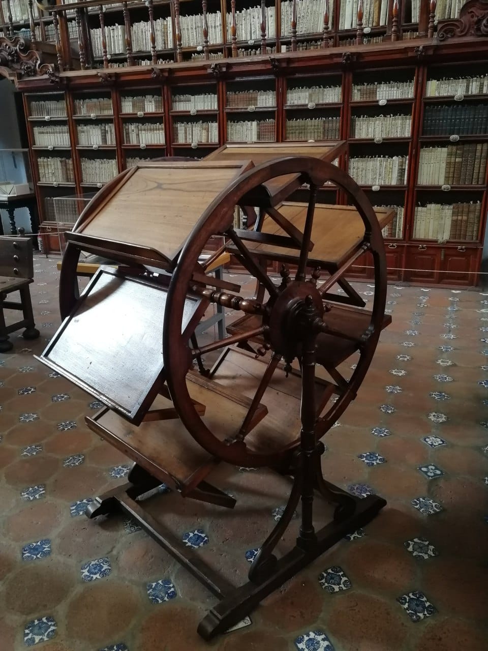 A wooden rotating wheel with tilted reading stands in an old library hall with bookcases and a tiled floor.