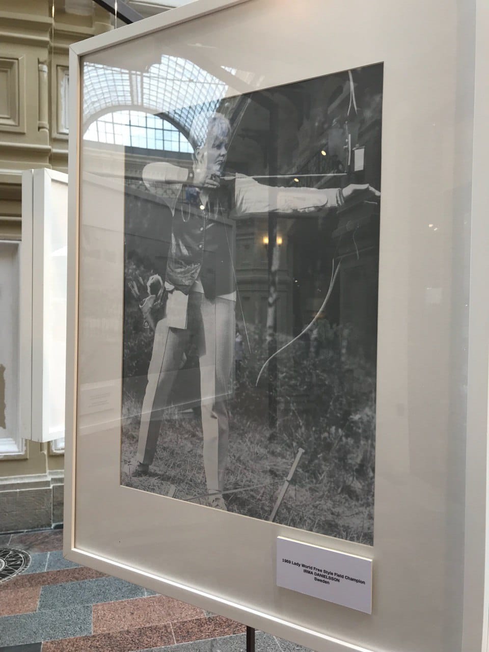 Photo en noir et blanc d'une archère bandant son arc, dans un cadre blanc sous verre. La légende indique Irma Danielsson, Suède, 1969. Une verrière apparaît derrière.