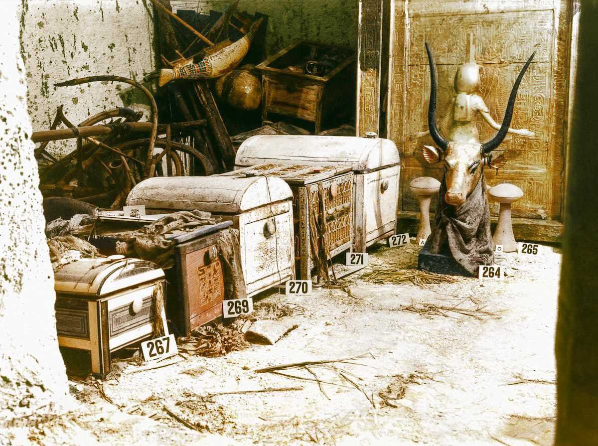 Des coffres blancs à couvercle bombé, numérotés, sur un sol poussiéreux devant une paroi dorée ; à droite, une tête de vache dorée aux cornes noires, au fond des objets en bois brisés.