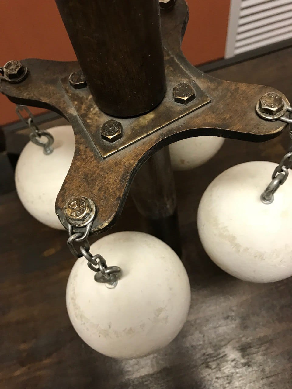 Un montant en bois est boulonné à une plaque métallique à quatre branches. Des boules blanches pendent à ses extrémités par des chaînes. Au fond, une table sombre et un mur orange.