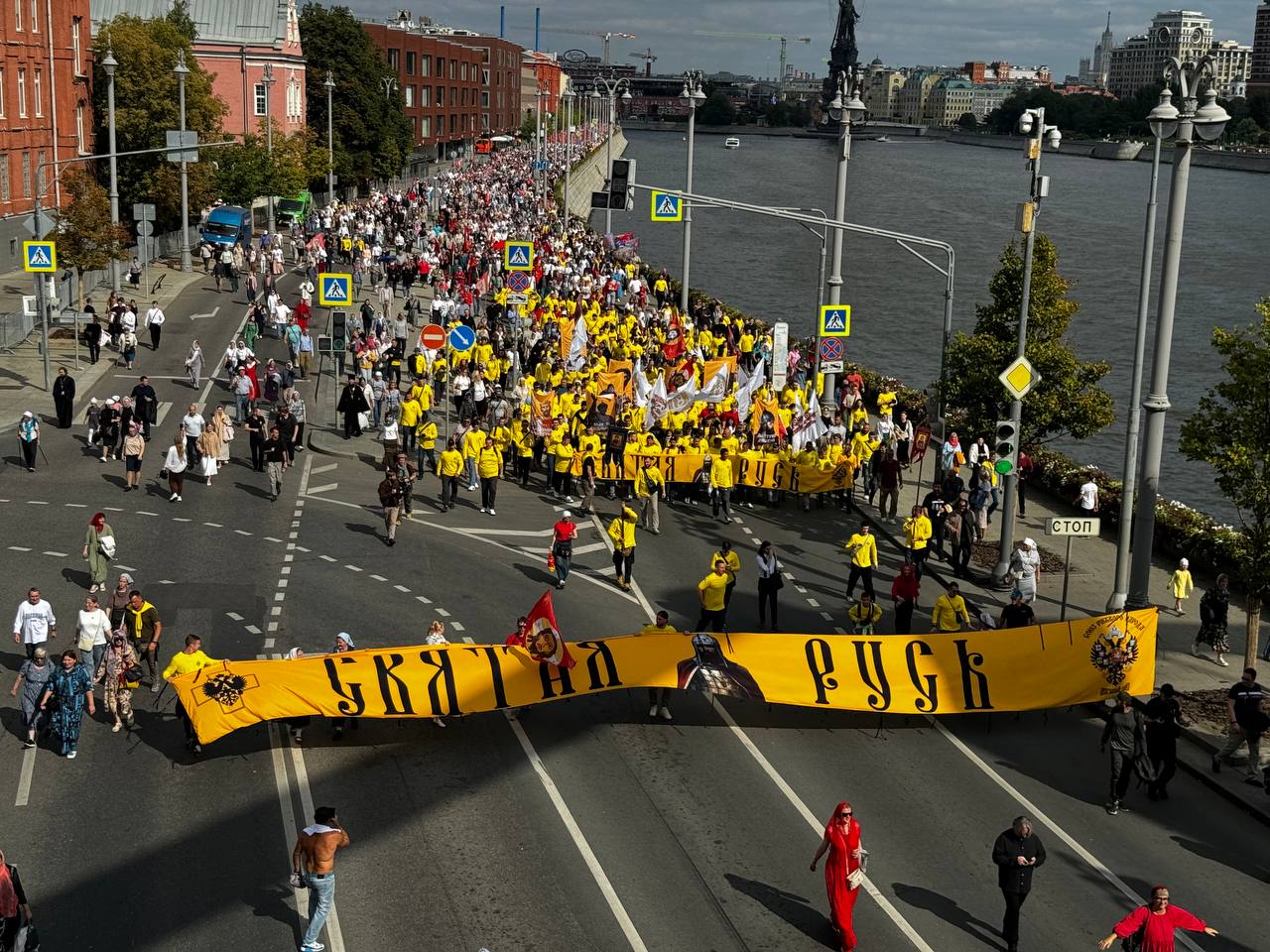 Aerial view of a column of people in yellow shirts walking along a riverside road with flags and banners. A yellow banner reading “Holy Rus” is stretched in front; the river is on the right.