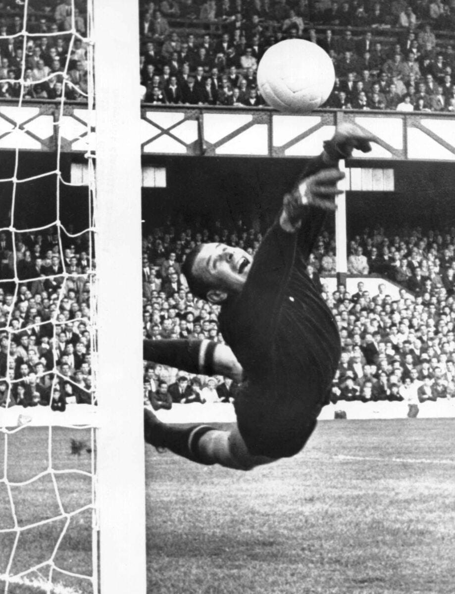 Black and white photo of a goalkeeper in dark kit diving to reach the ball beside the goalpost, with spectators in the stands behind.