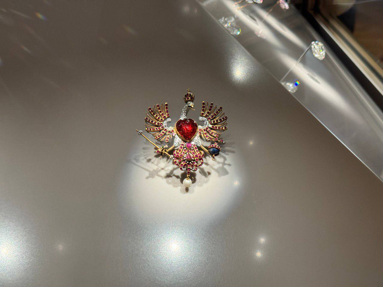 Витринадағы алтын broche: тәжді қыран, ұсақ рубиндермен көмкерілген қанаттар, кеудесінде жүрек пішінді ірі қызыл тас, скипетр, держава және салбыраған інжу.