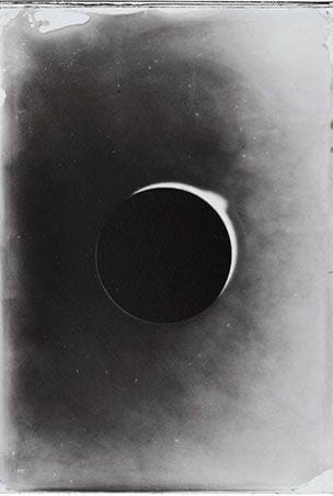 Black-and-white photograph of a dark disc with a thin bright crescent along its edge against a grey background.