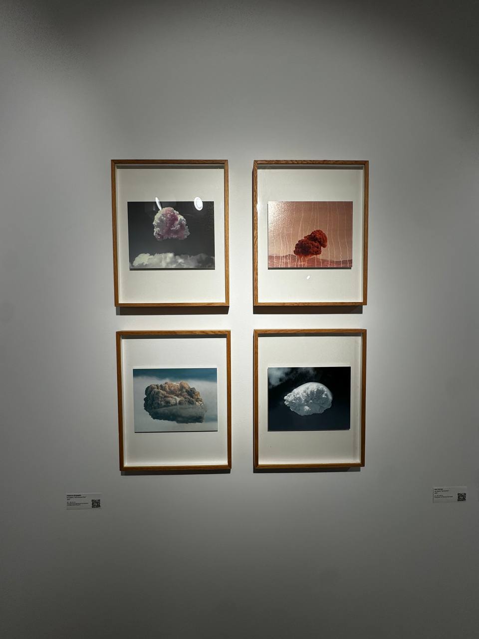 Four photographs in wooden frames hang on a pale wall in two rows of two. Each shows an isolated billowing cloud: pinkish, red, brownish and white. Labels with QR codes sit beside them.