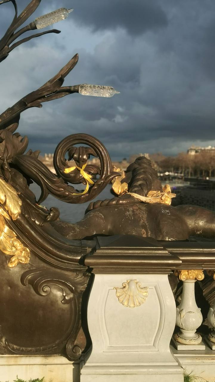 Bronze sculpture with gilded details, a scrolled railing and a glass lamp beside a marble pillar with a shell relief. Dark storm clouds and a river lie behind.