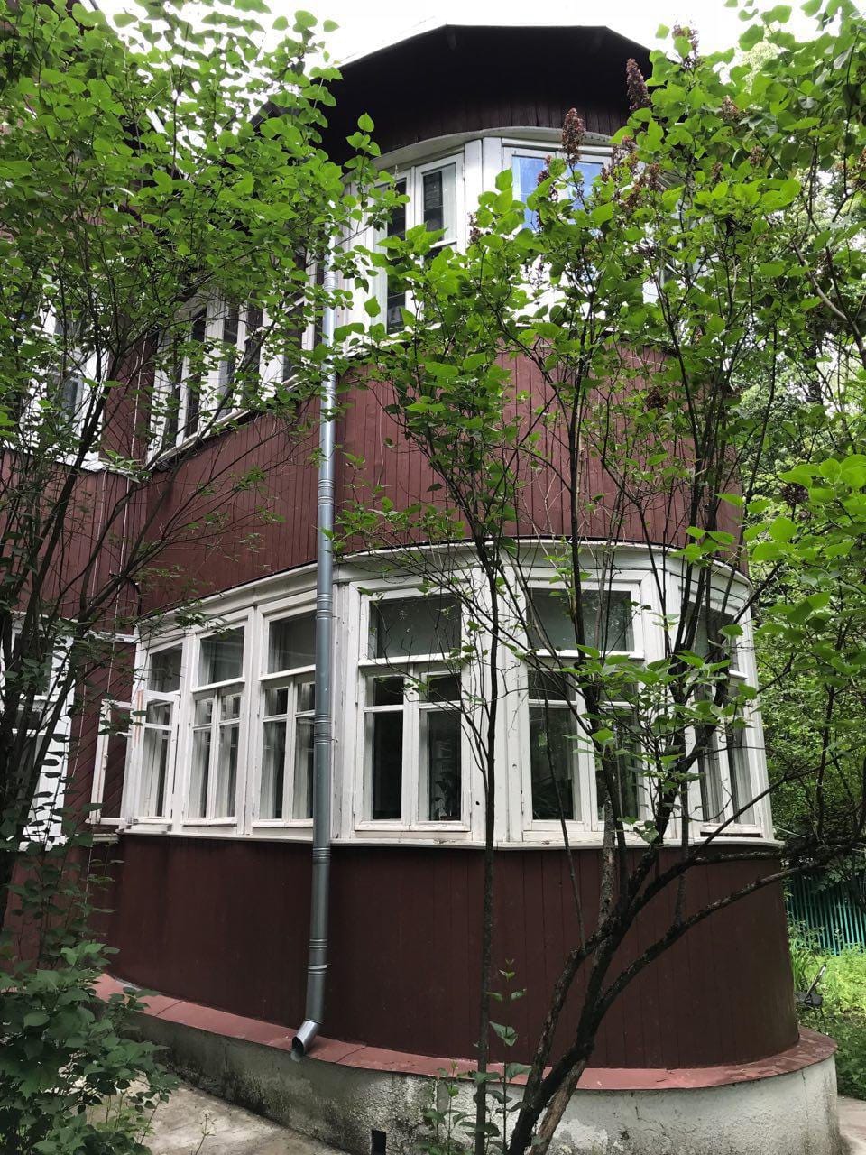 Maison en bois à deux étages, bardage rouge sombre et façade arrondie aux fenêtres blanches. Des branches de lilas devant, une gouttière le long du mur.