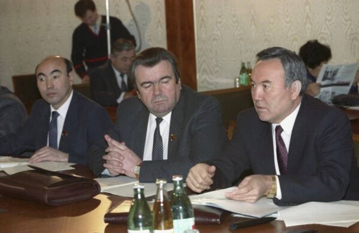 Three men in dark suits sit at a polished table with papers and bottles. The man on the right speaks and gestures; behind them people sit, one reading a newspaper.