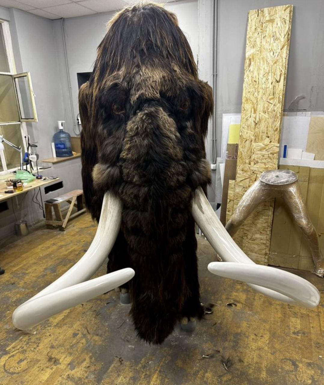 Front view of a mammoth figure with long dark fur and curved white tusks, standing in a workshop beside OSB boards and a worktable.