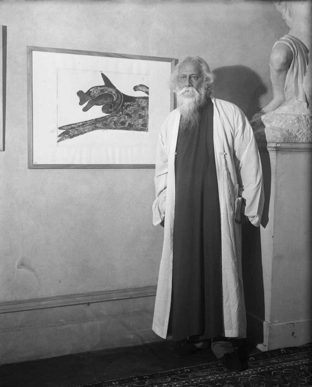 Black-and-white photo of a white-haired man with a long beard in a light long coat over a dark robe, standing by a framed drawing of a stylized animal. A sculpture is at right.