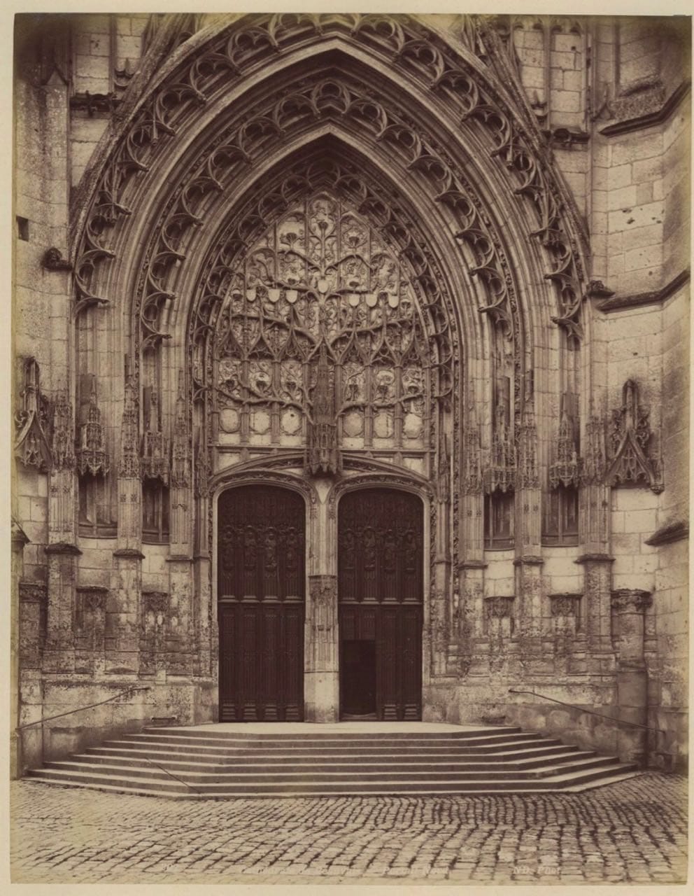 Ancienne photographie sépia d'un portail gothique à double porte, orné de sculptures, avec des marches devant l'entrée.