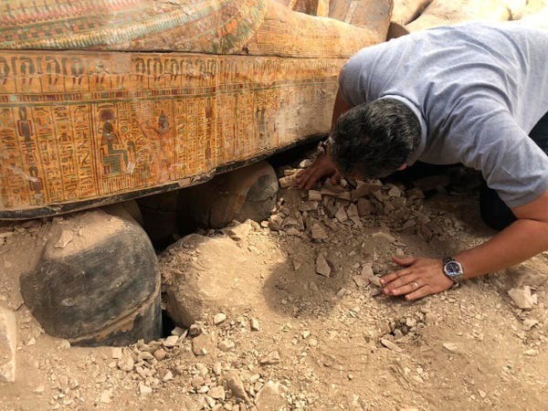 Un homme en T-shirt gris se penche près d'un sarcophage orné de hiéroglyphes et de figures, entouré de gravats et de pierres.